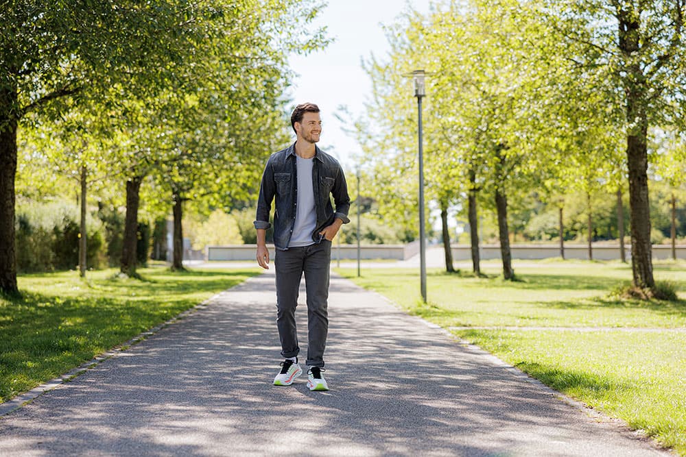 Ein Mann, der auf einem Parkweg mit Bäumen und Gras zu beiden Seiten spazieren geht und eine Jeansjacke und Jeans trägt.