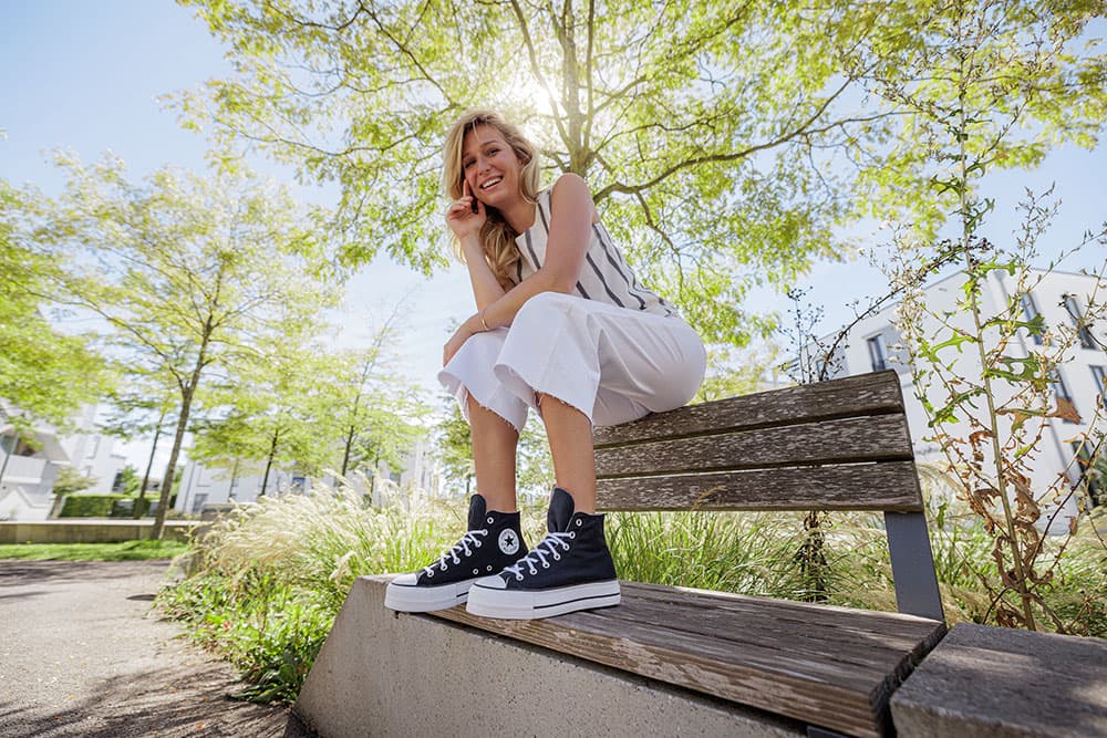 Eine Frau in einer weißen Hose und schwarzen Turnschuhen sitzt lächelnd auf einer Bank, im Hintergrund sind Bäume und ein strahlend blauer Himmel zu sehen.