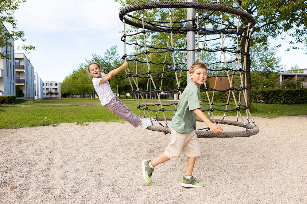 Zwei Kinder spielen auf einem Klettergerüst in einem sonnigen Park, im Hintergrund sind Gebäude und Bäume zu sehen.