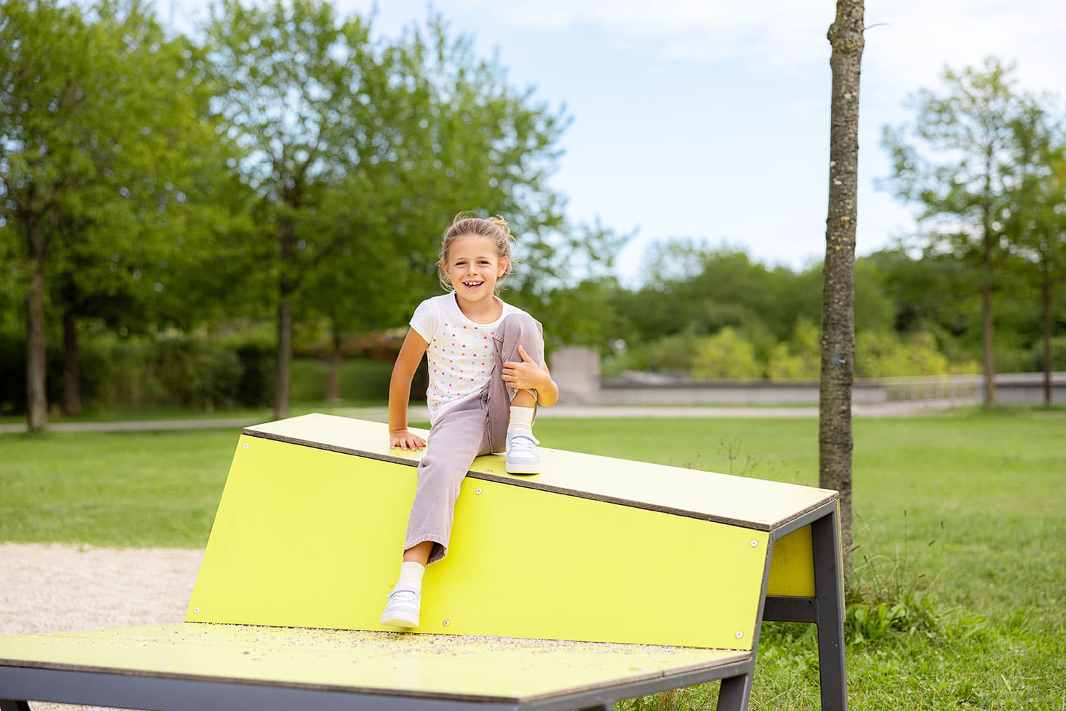 Ein junges Mädchen sitzt lächelnd auf einer gelben, schrägen Plattform in einem Park, umgeben von grünem Gras und Bäumen unter einem klaren Himmel.