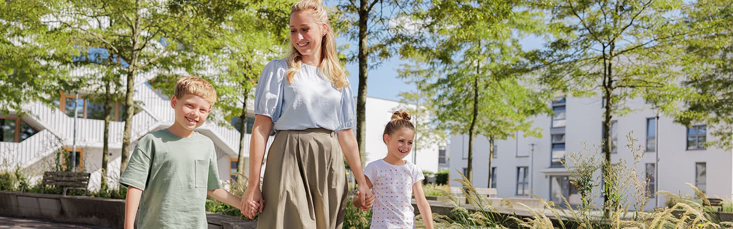 Eine Frau geht Hand in Hand mit einem lächelnden Jungen und Mädchen draußen spazieren, im Hintergrund moderne Gebäude und Bäume an einem sonnigen Tag.