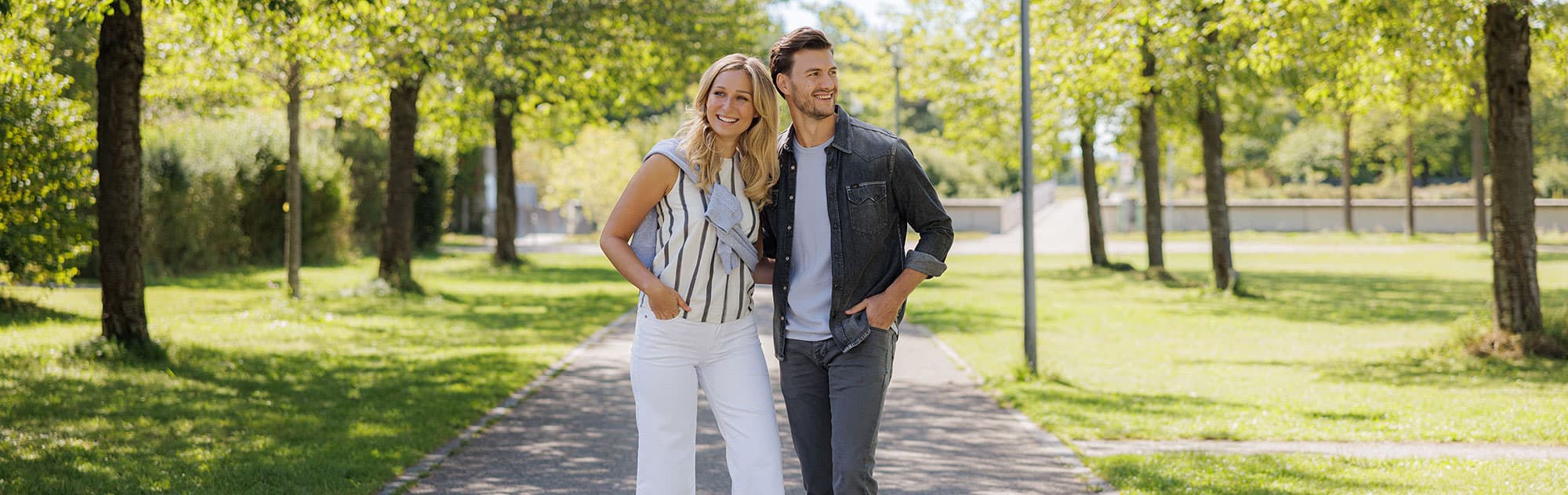 Ein lächelndes Paar spaziert auf einem Weg in einem Park, der von grünen Bäumen gesäumt ist.