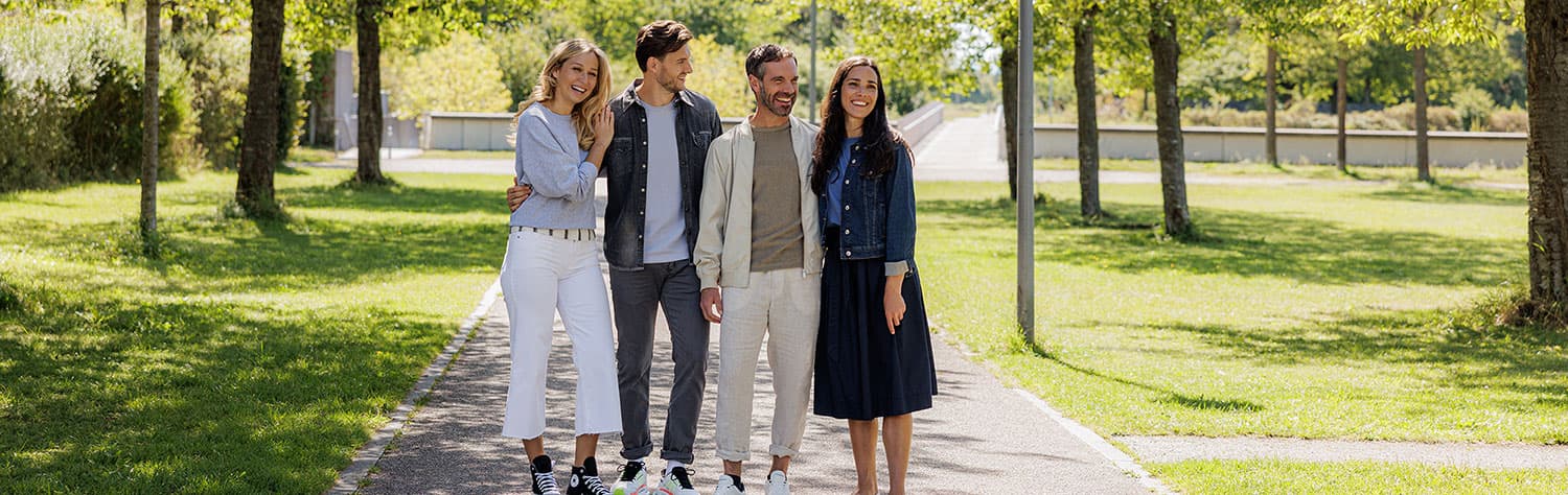 Vier Personen, die lächelnd in lässiger Frühlingskleidung auf einem von Bäumen gesäumten Weg in einem Park spazieren gehen.