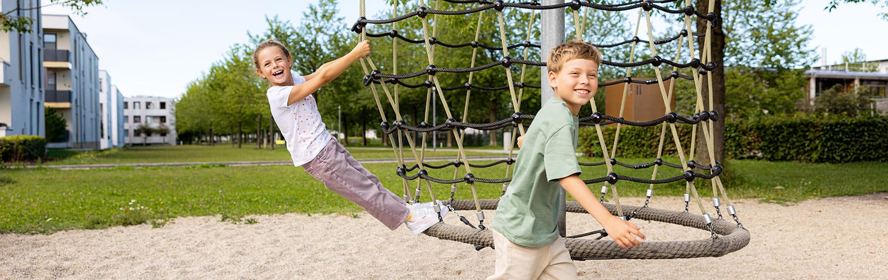 Zwei Kinder spielen fröhlich auf einer großen, runden Seilschaukel in einem Park, umgeben von Bäumen und Gebäuden.