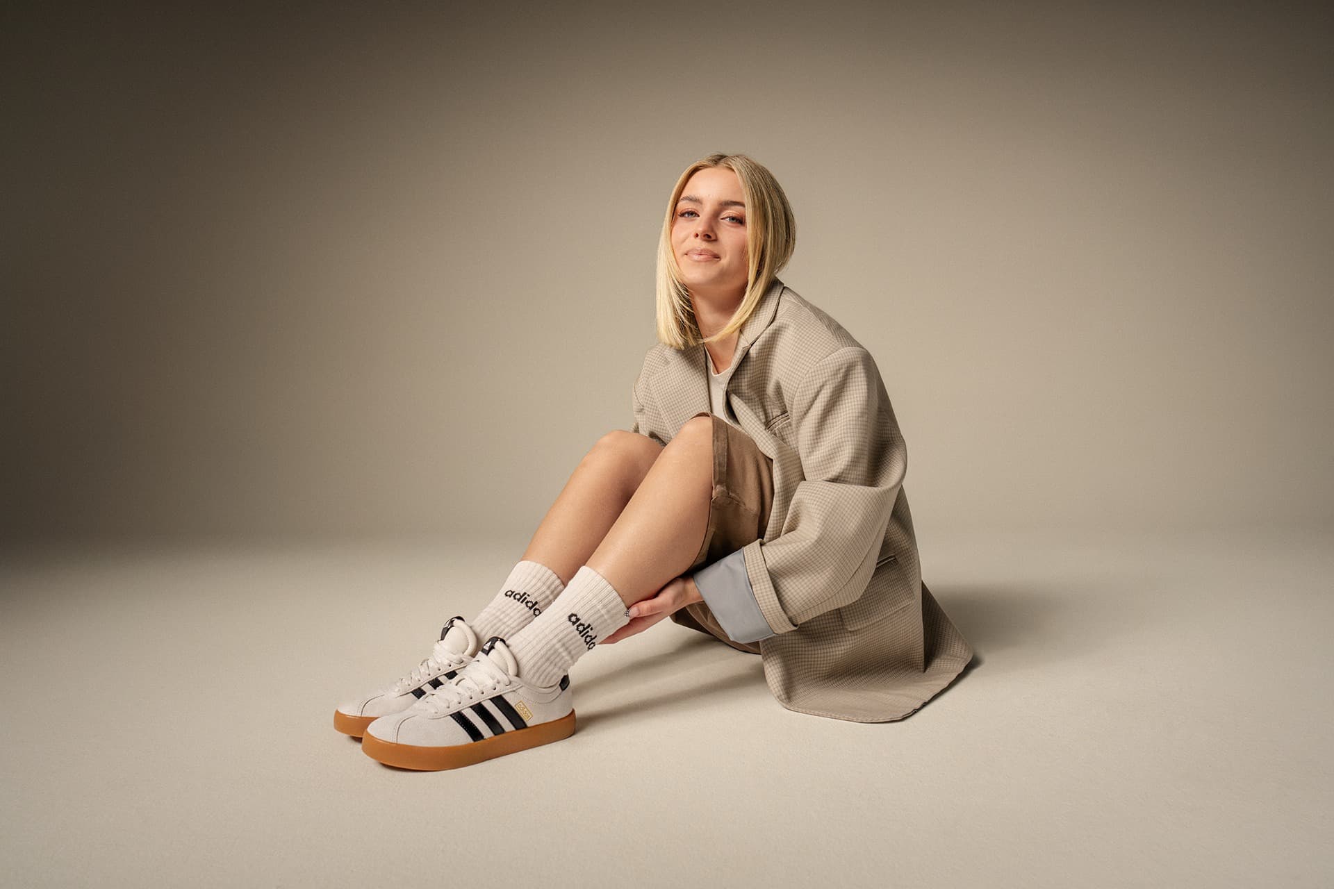 Eine Frau mit blonden Haaren sitzt auf dem Boden und trägt einen beigen Mantel, beige Socken und beige Turnschuhe mit schwarzen Streifen in einer neutralen Umgebung.