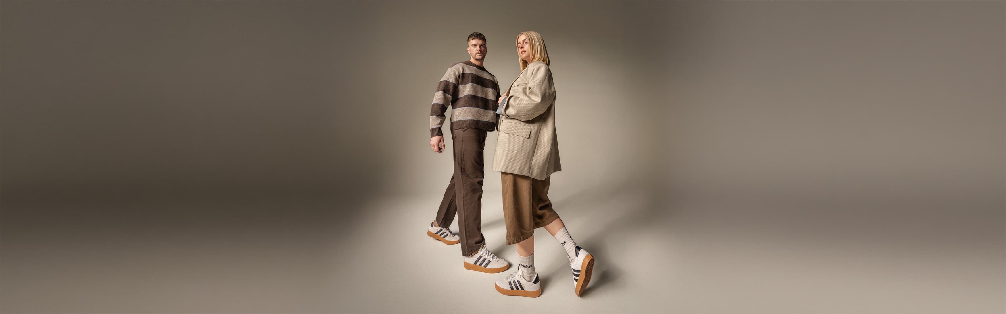 Ein Mann und eine Frau in Adidas-Turnschuhen und Freizeitkleidung stehen vor einem neutralen Hintergrund.